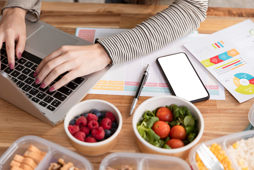 Imagem mostra mãos de mulher digitando em um computador e na mesa aparece potes com frutas e cereais