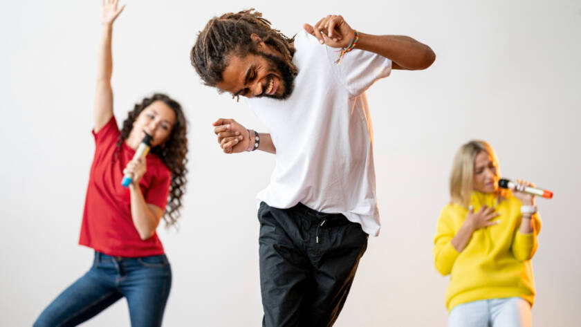 Homem dança enquanto duas mulheres cantam ao fundo.